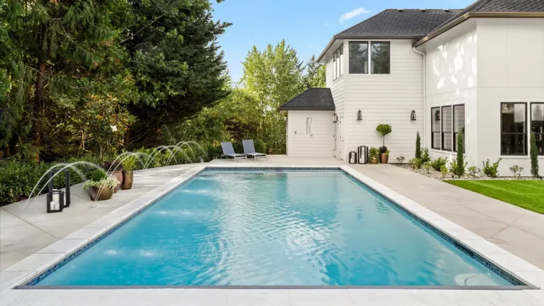 Beautiful home exterior and large swimming pool on sunny day with blue sky. Features series of water jets forming arches.